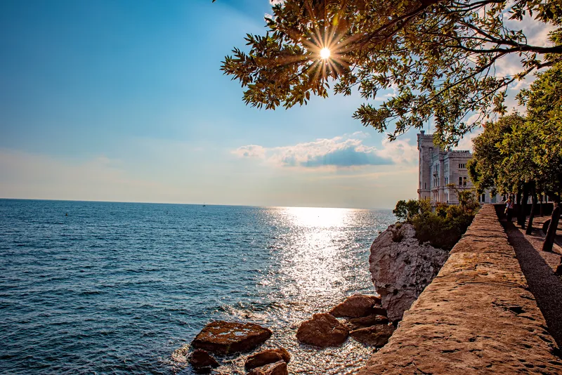 Il sole basso sul mare con gli scogli in primo piano e il castello sullo sfondo